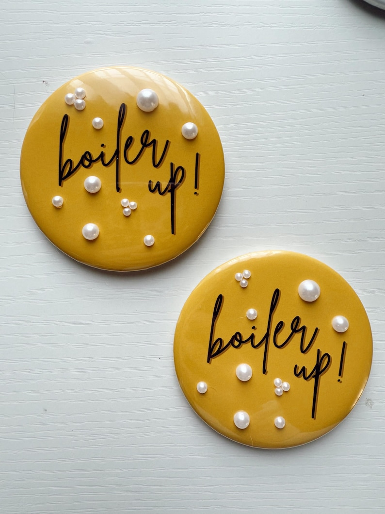 May include: Two round, mustard-yellow coasters with the phrase "boil er up!" in black script. Each coaster is adorned with faux pearls of varying sizes, adding a touch of elegance to the design. The coasters are placed on a white surface.