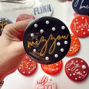 May include: A collection of round, glossy buttons with various phrases and designs. One button is black with "mizzou" in gold script and white pearl accents. Other buttons are red, white, and navy blue, with phrases like "go cards" and "roll tide".