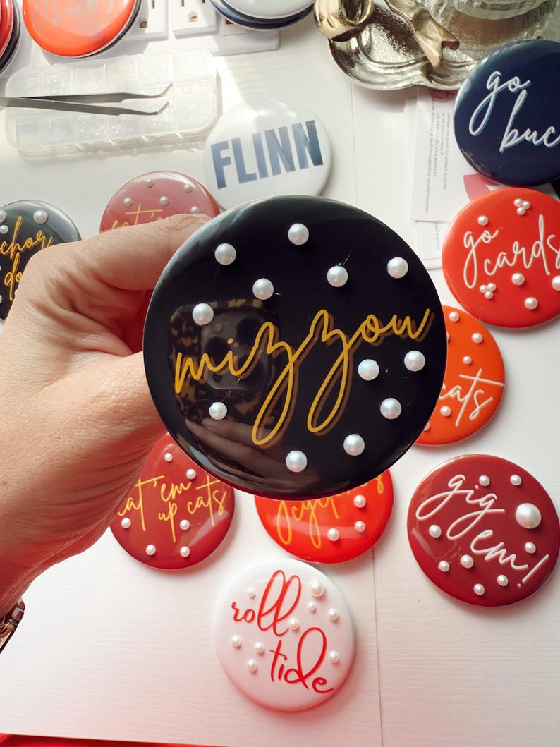 May include: A collection of round, glossy buttons with various phrases and designs. One button is black with "mizzou" in gold script and white pearl accents. Other buttons are red, white, and navy blue, with phrases like "go cards" and "roll tide".