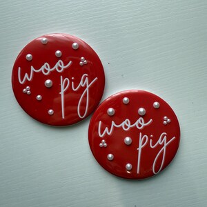 May include: Two red circular coasters with the words "woo pig" in white script. The coasters are decorated with small, white, pearl-like embellishments. The coasters are on a light blue surface.