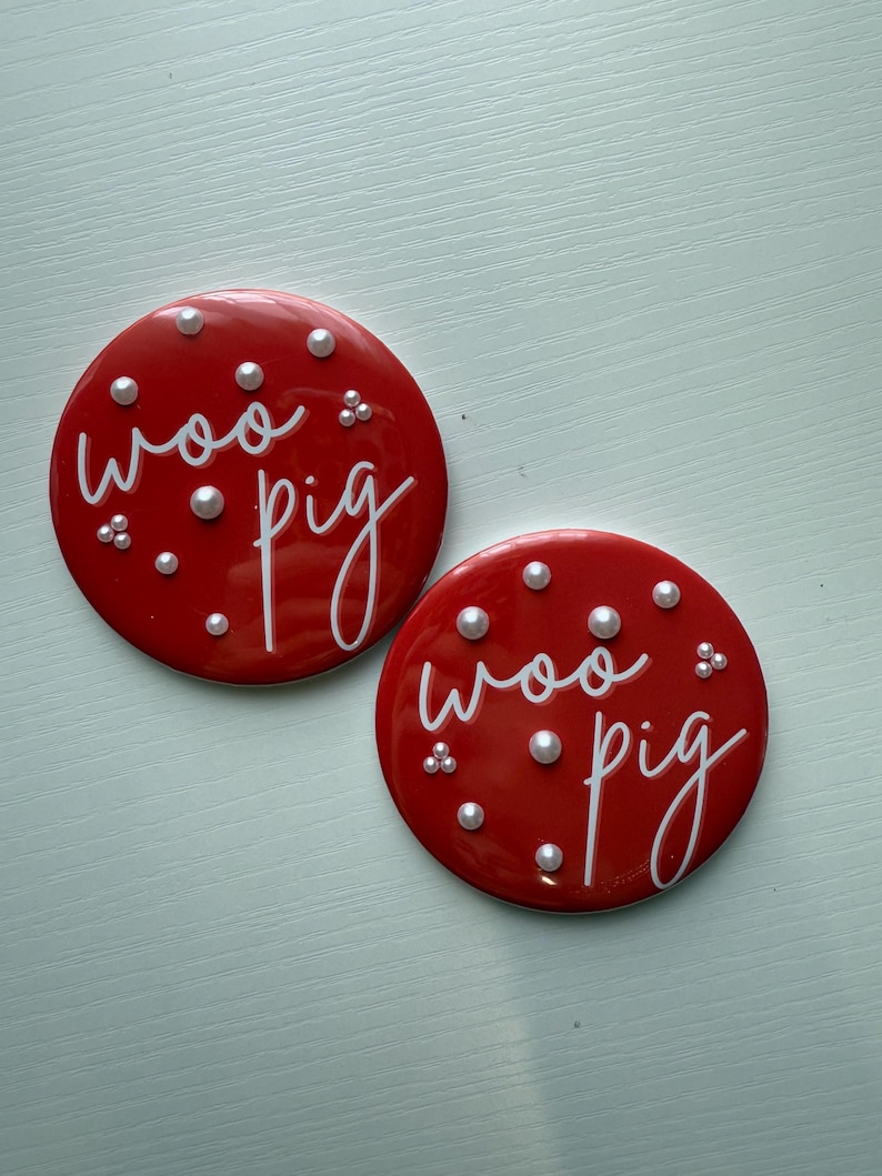 May include: Two red circular coasters with the words "woo pig" in white script. The coasters are decorated with small, white, pearl-like embellishments. The coasters are on a light blue surface.