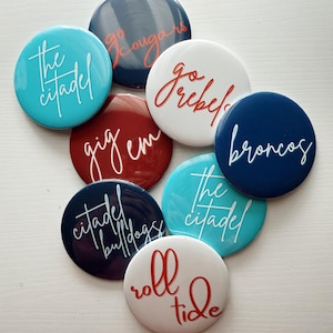 May include: A collection of round buttons in various colors, each featuring a different team name or slogan in a cursive font. Colors include teal, navy, red, and white. The buttons are arranged on a white surface.