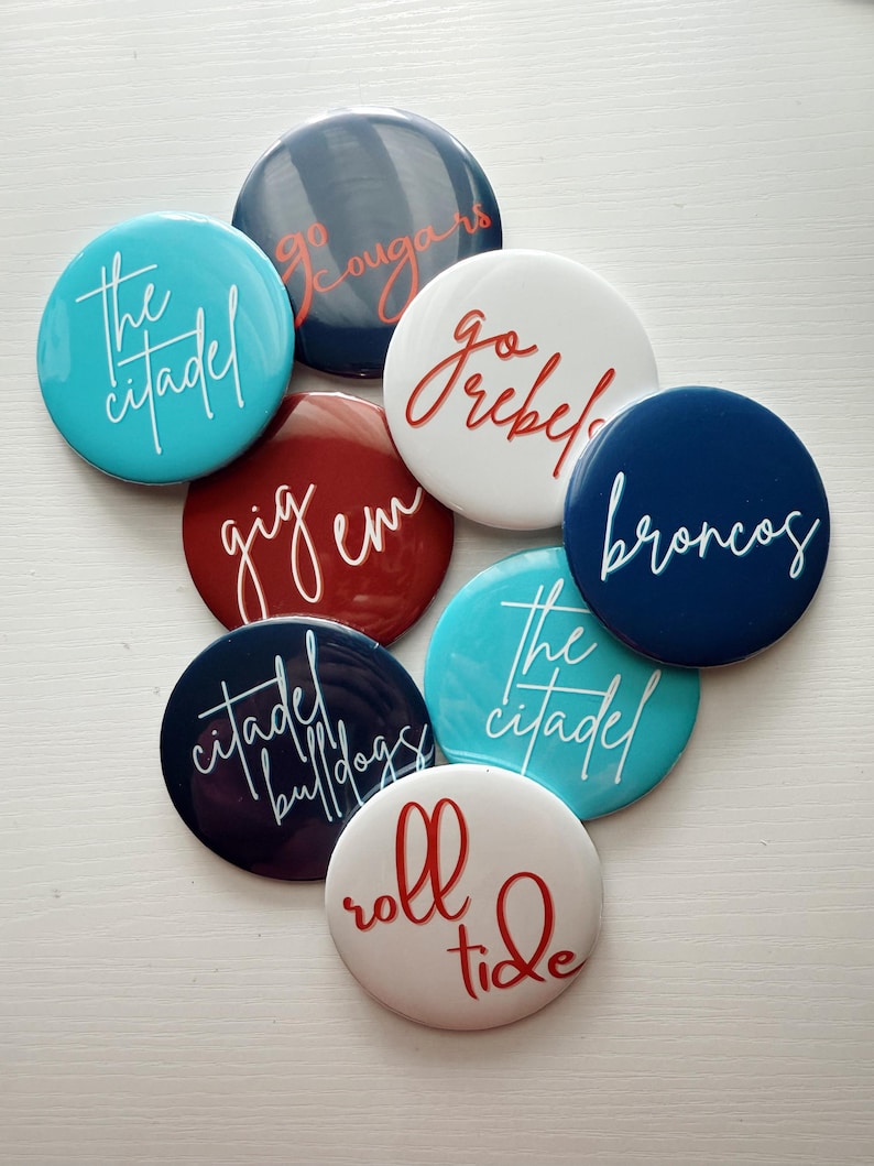 May include: A collection of round buttons in various colors, each featuring a different team name or slogan in a cursive font. Colors include teal, navy, red, and white. The buttons are arranged on a white surface.