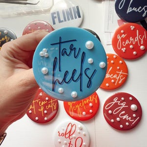 May include: A collection of round, colorful buttons with various team names and phrases. The buttons are in shades of blue, red, orange, and white, with white text and pearl accents. The buttons are a fun accessory for sports fans.