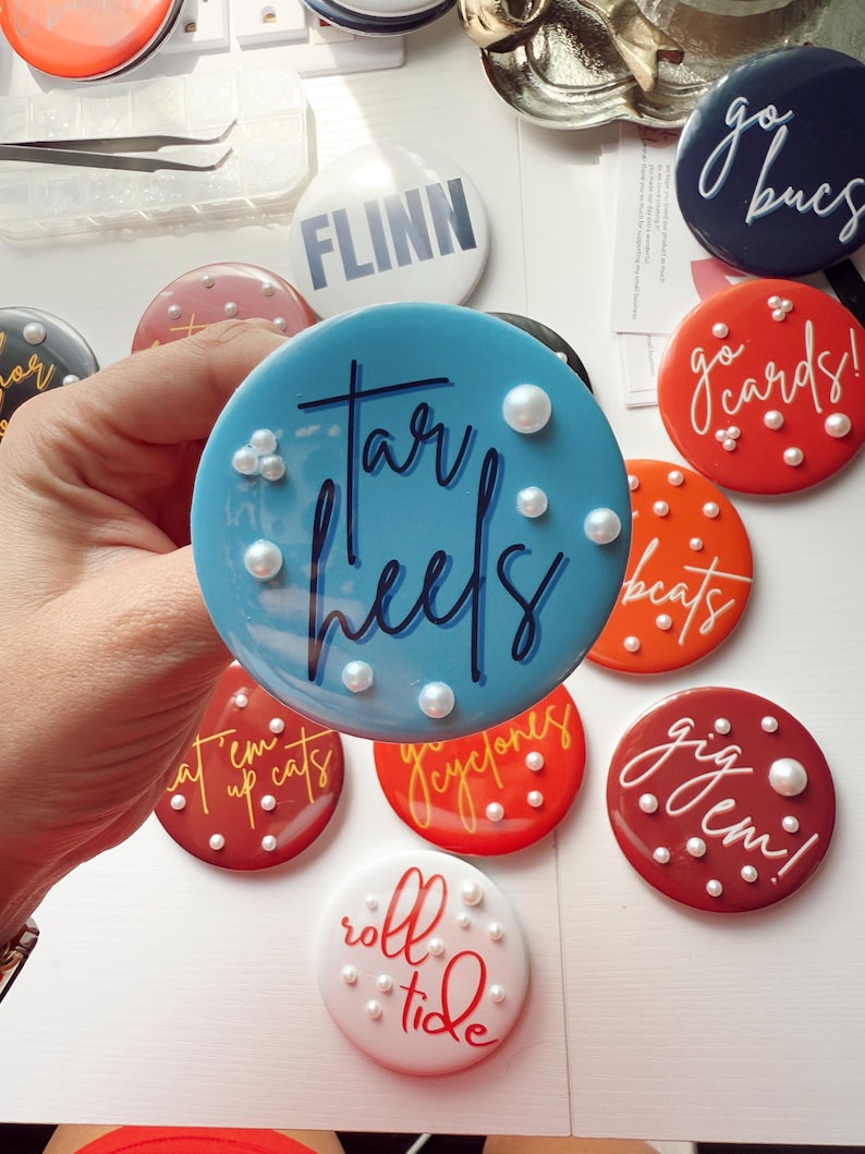 May include: A collection of round, colorful buttons with various team names and phrases. The buttons are in shades of blue, red, orange, and white, with white text and pearl accents. The buttons are a fun accessory for sports fans.