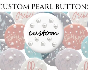 Peut inclure: Divers boutons de perles personnalisés dans des couleurs pastel, blanc, rose et bleu. Chaque bouton est orné de plusieurs perles et de texte personnalisé. Le bouton central affiche le mot "custom" en écriture noire.
