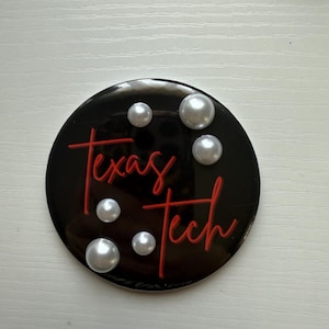 May include: A black round emblem with the words "Texas Tech" in red script. The emblem is decorated with six faux pearls. The background is a light, neutral color.