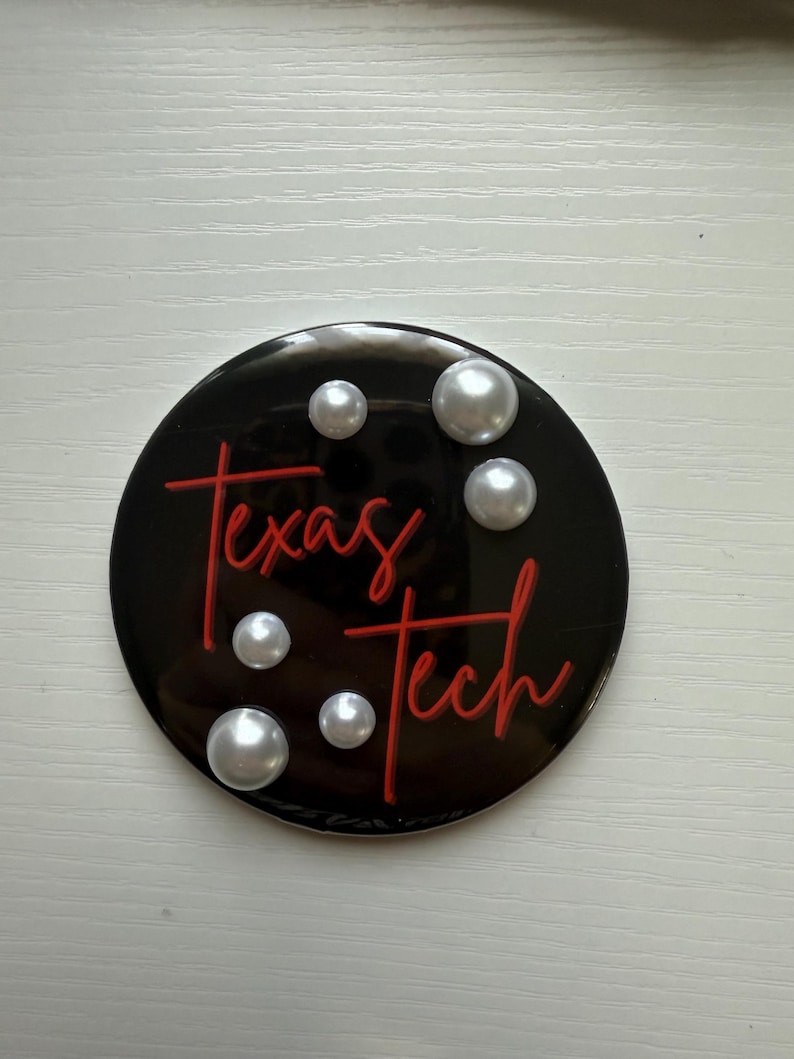 May include: A black round emblem with the words "Texas Tech" in red script. The emblem is decorated with six faux pearls. The background is a light, neutral color.