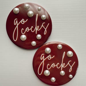 May include: Two round, maroon coasters with the words "go cocks" in white script. Each coaster is adorned with several white, pearl-like embellishments. The coasters are displayed on a white surface.