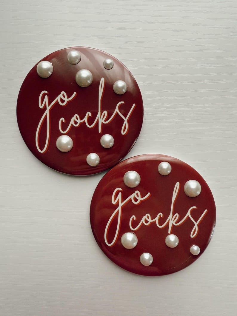 May include: Two round, maroon coasters with the words "go cocks" in white script. Each coaster is adorned with several white, pearl-like embellishments. The coasters are displayed on a white surface.