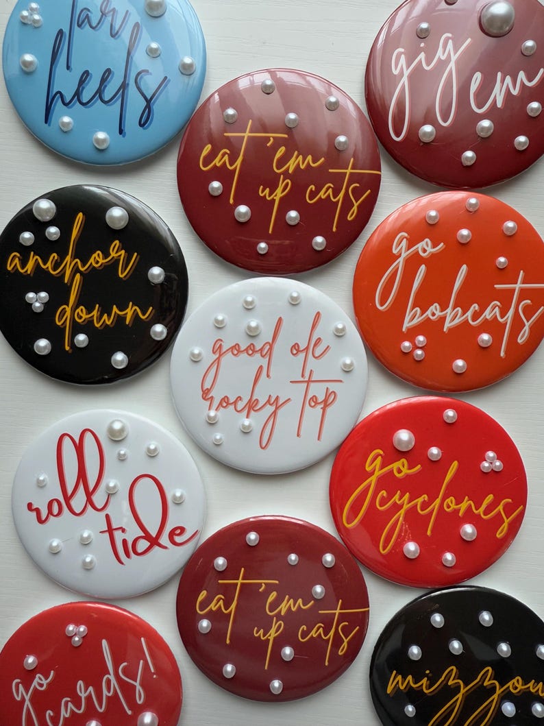 May include: A collection of round, glossy buttons in various colors, each featuring a different phrase in cursive script. The buttons are adorned with small, pearl-like embellishments. Phrases include "Tar Heels," "Anchor Down," and "Roll Tide."