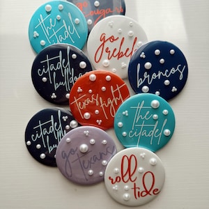 May include: A collection of round, colorful coasters with various team names and phrases in cursive script. Each coaster is adorned with small, pearl-like embellishments. Colors include teal, navy, red, white, and purple.