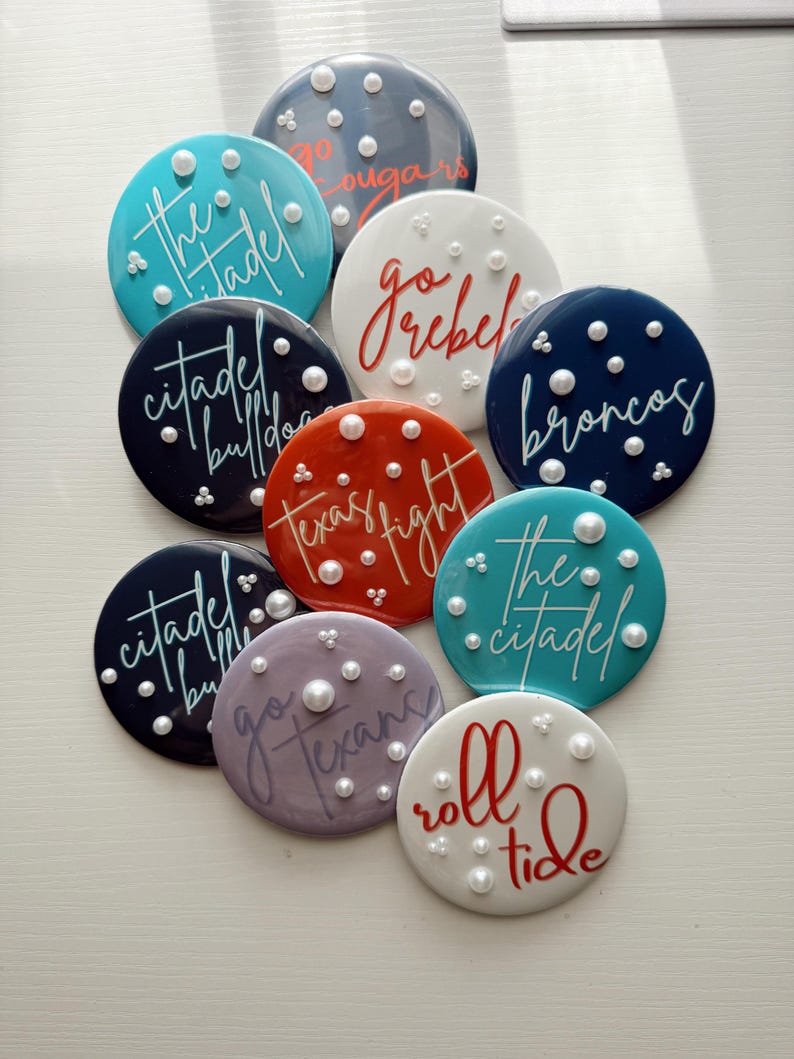 May include: A collection of round, colorful coasters with various team names and phrases in cursive script. Each coaster is adorned with small, pearl-like embellishments. Colors include teal, navy, red, white, and purple.