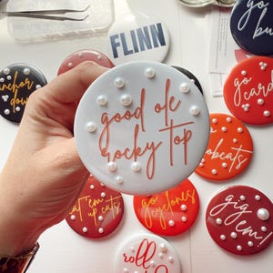 May include: A collection of round, decorative buttons in various colors, featuring phrases like "good ole rocky top" and "roll tide." Each button is adorned with small, pearl-like embellishments. Some buttons have team names or logos.