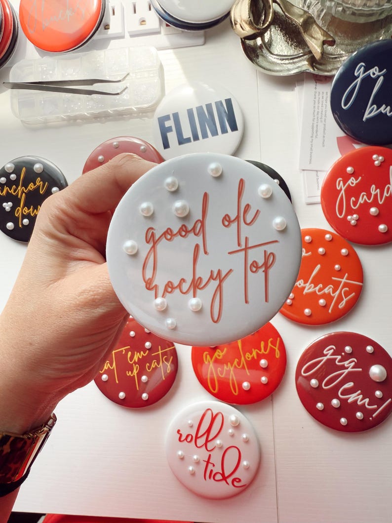 May include: A collection of round, decorative buttons in various colors, featuring phrases like "good ole rocky top" and "roll tide." Each button is adorned with small, pearl-like embellishments. Some buttons have team names or logos.