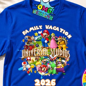 May include: A royal blue short-sleeve t-shirt featuring a colorful graphic of various Nintendo characters, the text "FAMILY VACATION UNIVERSAL STUDIOS" in a curved font, and the year "2026" printed in white at the bottom center of the design.