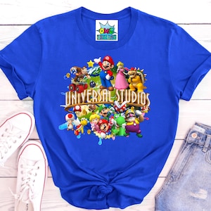 May include: A royal blue t-shirt featuring a graphic print of various Nintendo characters including Mario, Luigi, Peach, and Bowser, with the text Universal Studios centered in gold lettering. The shirt has short sleeves and a crew neckline.