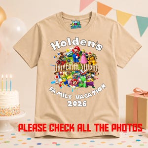 May include: A tan short-sleeved t-shirt featuring a colorful graphic of various Nintendo characters, the text "Holden's UNIVERSAL STUDIOS FAMILY VACATION 2026", and a small logo on the inner neck label. The shirt is displayed against a festive party background.