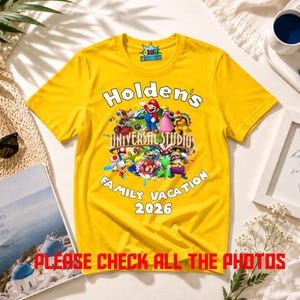 May include: A bright yellow short-sleeve t-shirt featuring a graphic of various Nintendo characters over a globe. The text reads Holden's UNIVERSAL STUDIOS FAMILY VACATION 2026. The shirt has a round neckline and a small logo on the inner collar.