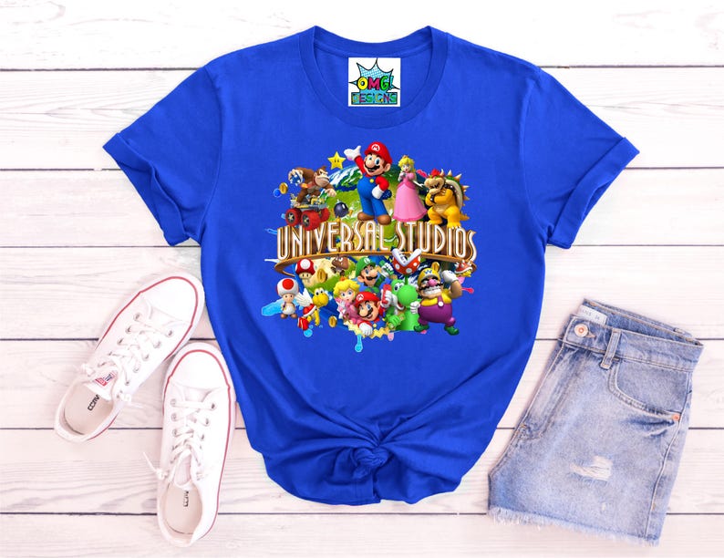 May include: A royal blue t-shirt featuring a graphic print of various Nintendo characters including Mario, Luigi, Peach, and Bowser, with the text Universal Studios centered in gold lettering. The shirt has short sleeves and a crew neckline.