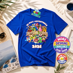 May include: A royal blue short-sleeve t-shirt featuring a colorful graphic of various Nintendo characters, the text "FAMILY ADVENTURES UNIVERSAL STUDIOS" and "2026". The shirt also displays a small "OMG! DESIGNS" logo at the top center.