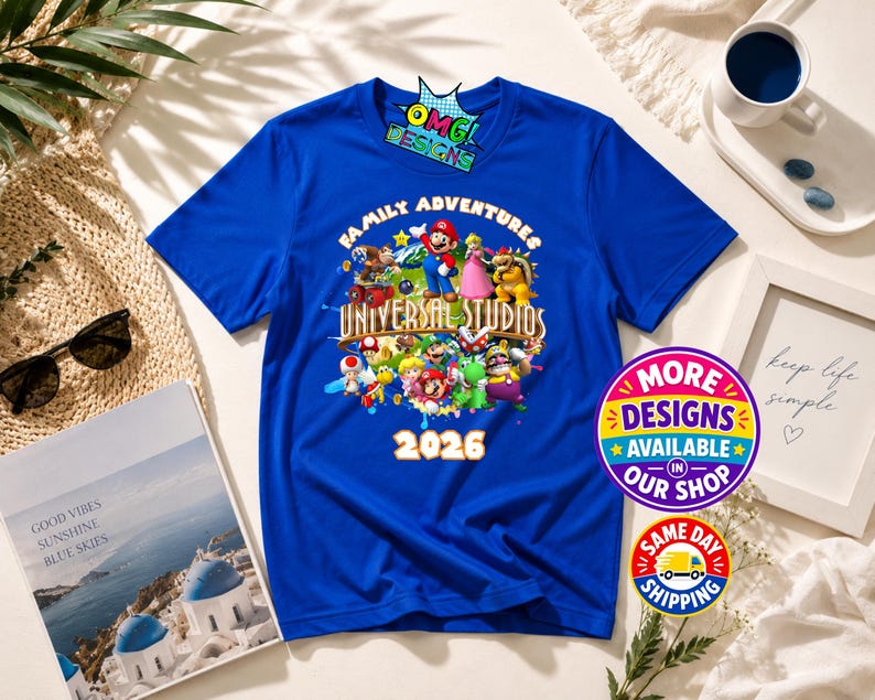 May include: A royal blue short-sleeve t-shirt featuring a colorful graphic of various Nintendo characters, the text "FAMILY ADVENTURES UNIVERSAL STUDIOS" and "2026". The shirt also displays a small "OMG! DESIGNS" logo at the top center.