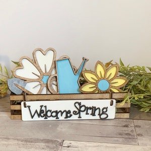 May include: Wooden crate with a "Welcome Spring" sign and spring-themed decorations, including a blue watering can, a yellow flower, and two white flowers.
