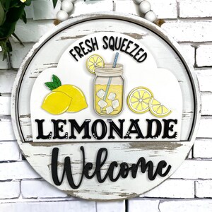 May include: A round wooden sign with a white background and black lettering that reads "FRESH SQUEEZED LEMONADE Welcome." The sign features a lemon, a glass of lemonade, and lemon slices. The sign is decorated with a string of white beads.