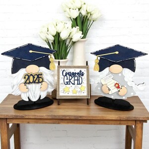 May include: Two graduation gnome decorations with black hats and white beards. One gnome holds a diploma and the other has the year 2025 on its chest. A wooden sign with the text "Congrats Grad" and three gold stars is in the center.