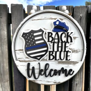 May include: A white wooden round sign with a black and blue police badge, a blue police hat, and the text "Back the Blue" and "Welcome".