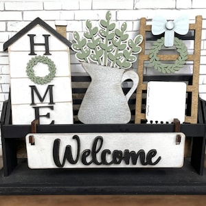 May include: A black wooden shelf with a white sign that says "Welcome". The shelf also has a white house with the word "HOME" on it, a white pitcher with green leaves, and a wooden ladder with a white bow and a green wreath.