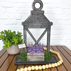 May include: A decorative wooden lantern with a weathered gray finish. It features a wooden box with purple lavender and green leaves. A sign reads "Fresh Lavender 25¢". A wooden bead garland is draped around the base.