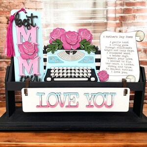 May include: A black wooden display with a light blue typewriter, pink roses, and a scroll with a poem. The display also features a sign that says "LOVE YOU" and a bookmark that says "best MOM ever".