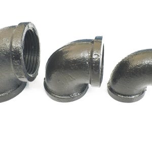 May include: Three black iron pipe fittings, each with a 90-degree angle, are shown on a white background. The fittings are threaded on one end and have a smooth, black finish.