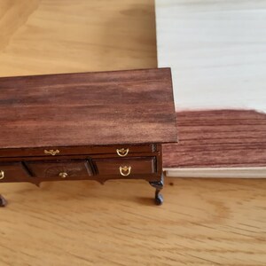May include: A dark brown wooden dresser with four drawers and gold-colored handles. The dresser has four legs and is sitting on a light brown wooden surface. A sample of dark brown wood stain is visible on the right side of the image.