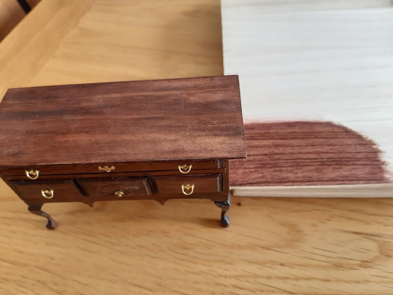 May include: A dark brown wooden dresser with four drawers and gold-colored handles. The dresser has four legs and is sitting on a light brown wooden surface. A sample of dark brown wood stain is visible on the right side of the image.