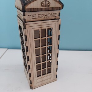 May include: A wooden model of a classic red British telephone booth with a crown on top and the word "TELEPHONE" printed on the front.