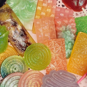 May include: A collection of colorful, glittery, and decorative soaps in various shapes, including rectangles, circles, and flower-like designs. The soaps are in shades of green, orange, pink, and purple, arranged on a pink lace doily.