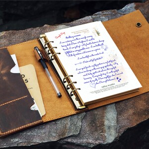May include: Open leather journal with a pen, a notepad, and a card holder. The journal is brown with a lighter tan interior and gold-colored rings. The notepad has handwritten text in blue ink. A black pen rests on the open pages.