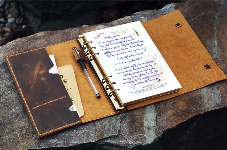 May include: Open leather journal with a pen, a notepad, and a card holder. The journal is brown with a lighter tan interior and gold-colored rings. The notepad has handwritten text in blue ink. A black pen rests on the open pages.
