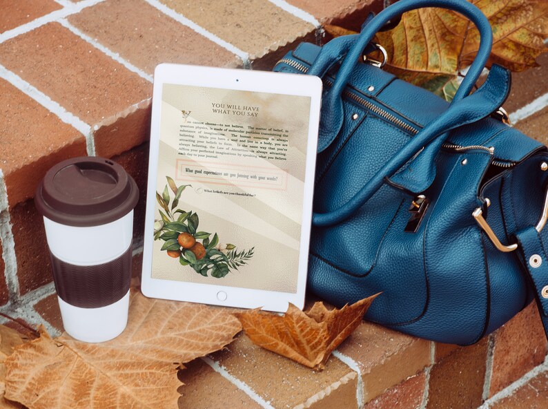 May include: A white tablet displays text and botanical illustrations, next to a teal handbag and a white travel mug with a brown lid. The tablet's screen features the words "YOU WILL HAVE WHAT YOU SAY". The scene is set on a brick surface with fallen leaves.