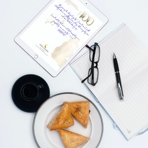 May include: An overhead shot of a workspace with a tablet displaying text, a notebook, glasses, a pen, a cup of coffee, and a plate with three pastries. The tablet has the text "The journey has changed my life! I can't wait... GRATEFUL!"