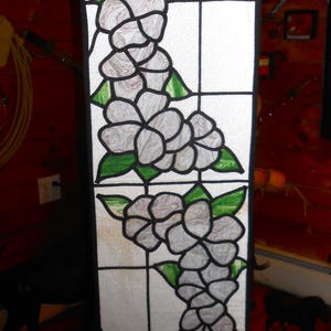 May include: A stained glass window panel with a white background and black grid lines. The panel features a design of white flowers with green leaves.