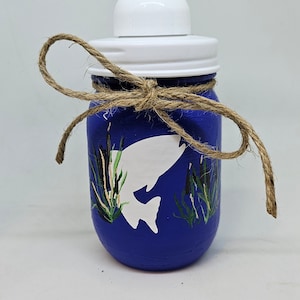 May include: Blue glass soap dispenser with a white pump and a brown twine bow. The dispenser is decorated with a white silhouette of a fish and green and black painted reeds.