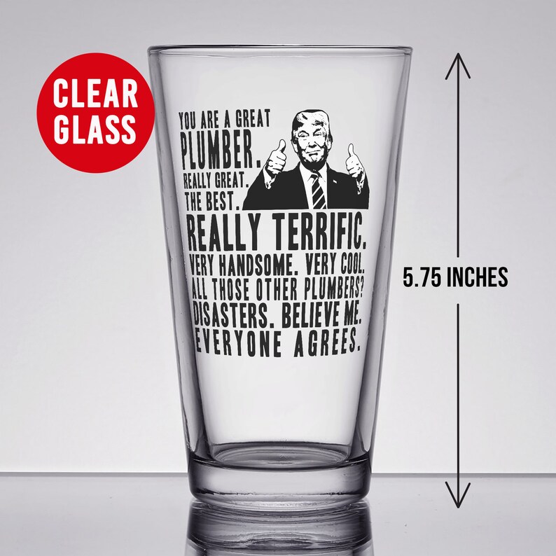 May include: A clear glass pint glass with a black and white image of a man with the text "You are a great plumber. Really great. The best. Really terrific. Very handsome. Very cool. All those other plumbers? Disasters. Believe me. Everyone agrees."