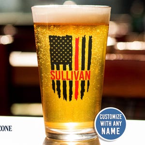 May include: A clear glass pint beer mug filled with golden liquid. The front features a black and red graphic of a distressed American flag with a thin red line and the name SULLIVAN printed in red. A blue circular badge reads CUSTOMIZE WITH ANY NAME.