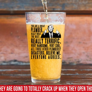 May include: A glass beer stein with a humorous graphic of a man with a thumbs up gesture and the text "You are a great plumber. Really great. The best. Really terrific. Very handsome. Very cool. All those other plumbers? Disasters. Believe me. Everyone agrees."