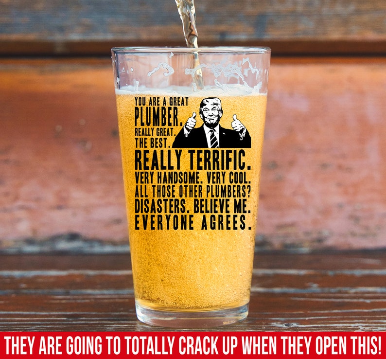 May include: A glass beer stein with a humorous graphic of a man with a thumbs up gesture and the text "You are a great plumber. Really great. The best. Really terrific. Very handsome. Very cool. All those other plumbers? Disasters. Believe me. Everyone agrees."