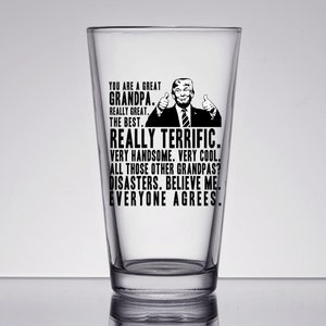 May include: A clear glass pint glass with a black and white image of a man with a thumbs up gesture and the text "You are a great Grandpa. Really great. The best. Really terrific. Very handsome. Very cool. All those other Grandpas? Disasters. Believe me. Everyone agrees."