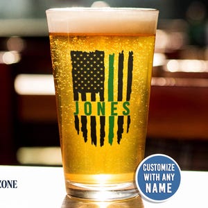 May include: A clear glass pint glass filled with golden beer, featuring a black and green distressed American flag graphic with the name JONES printed in green across the center. A blue circular badge reads CUSTOMIZE WITH ANY NAME.